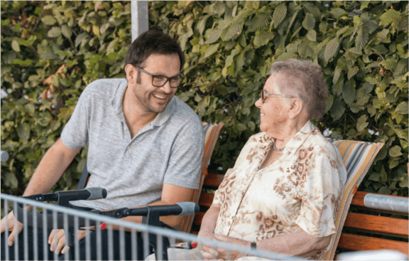 Betreuer und ältere Dame unterhalten sich auf einer Bank sitzend